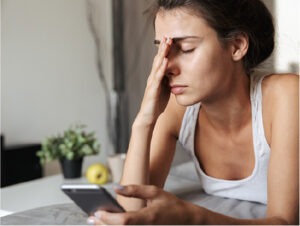 A tired and frustrated woman rubbing her eyes as she takes a break from looking at her phone.