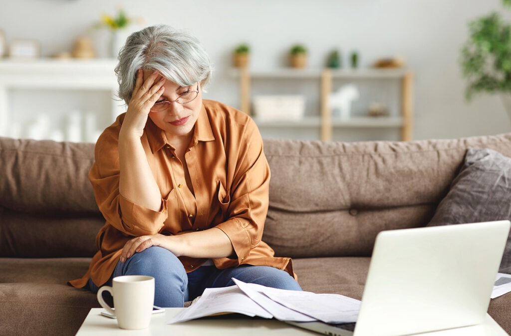 A stressed senior woman staring at piles of paper next to her laptop.